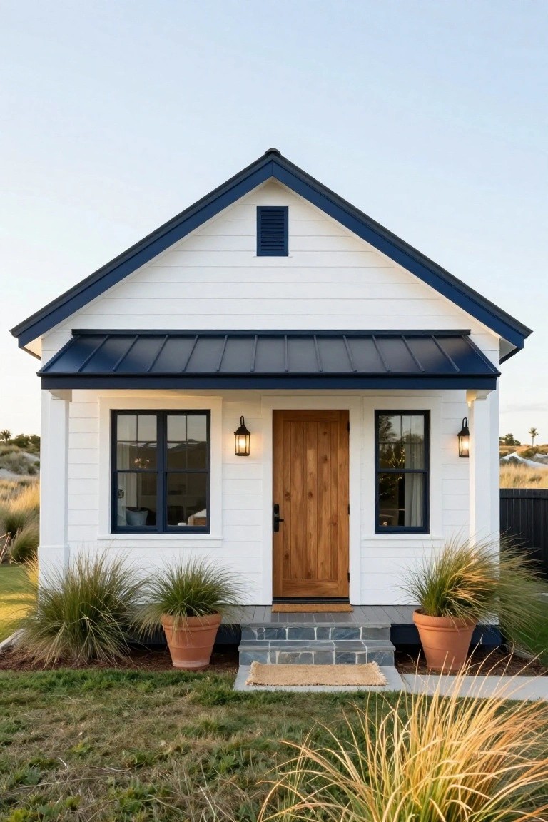Small white clapboard house with navy blue standing-seam metal roof, porch awning, window trim, wooden front door, lanterns, and potted grasses on stone steps amid lawn and dune grasses.
