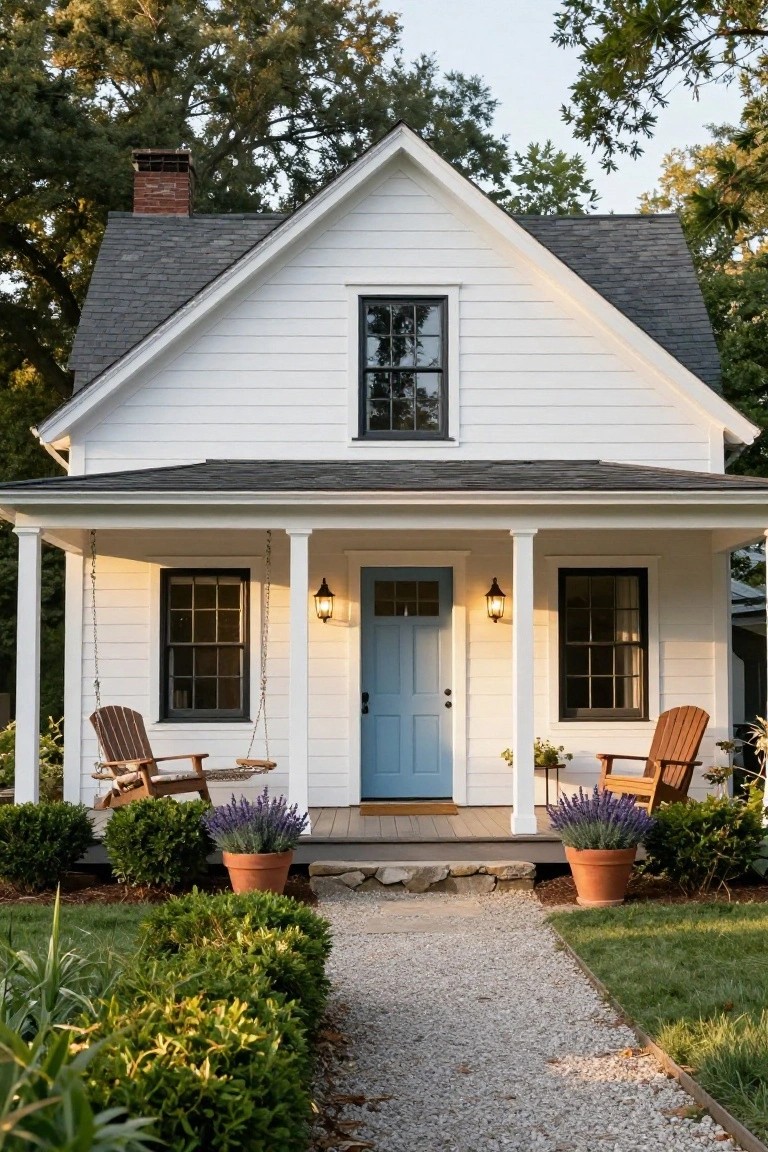White clapboard farmhouse with gabled roof, blue front door, black-framed windows, covered porch with Adirondack chairs and potted lavender, gravel path, boxwoods, and lawn.