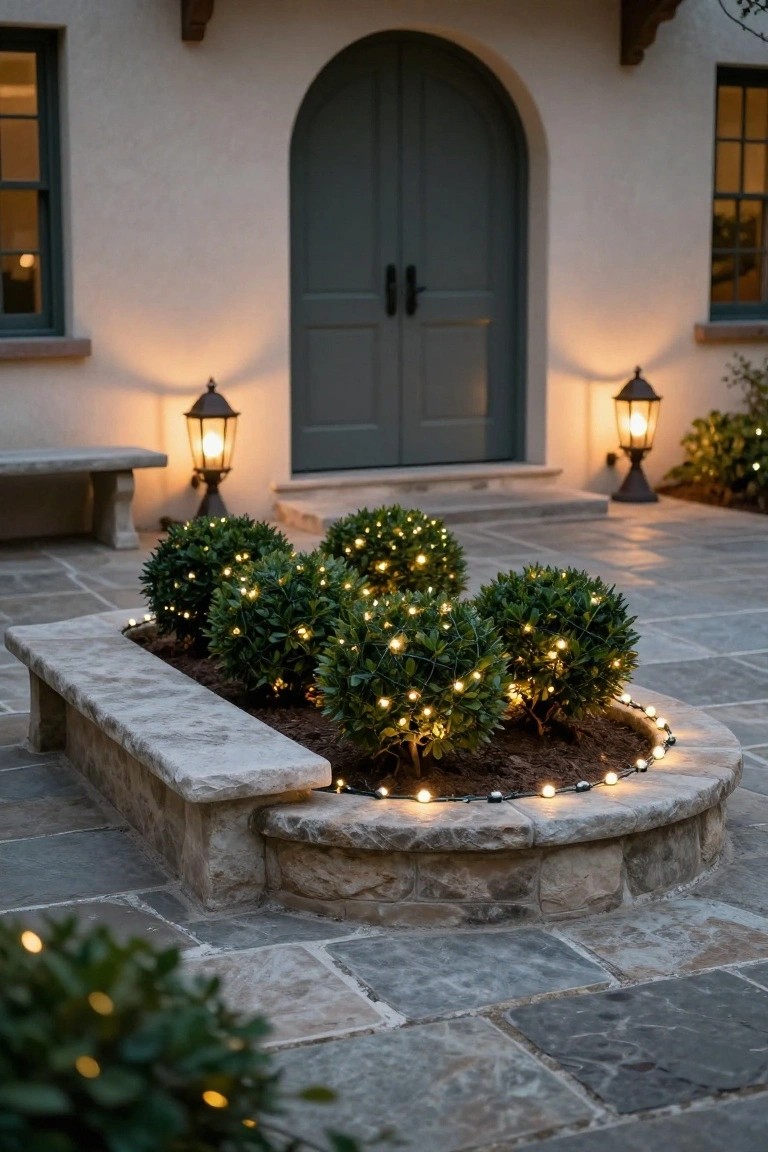 Arched green door on stucco house entrance with lanterns on each side, curved stone planter bed containing multiple round boxwood shrubs wrapped in white string lights, stone paver patio at dusk.