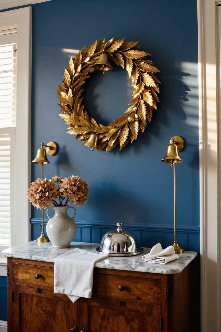 Gold laurel leaf wreath with brass bell on navy blue wall above marble-topped wooden console table with cloche, white napkins, and dried hydrangea bouquet in white vase, flanked by tall gold bell sconces and shorter wall sconces.