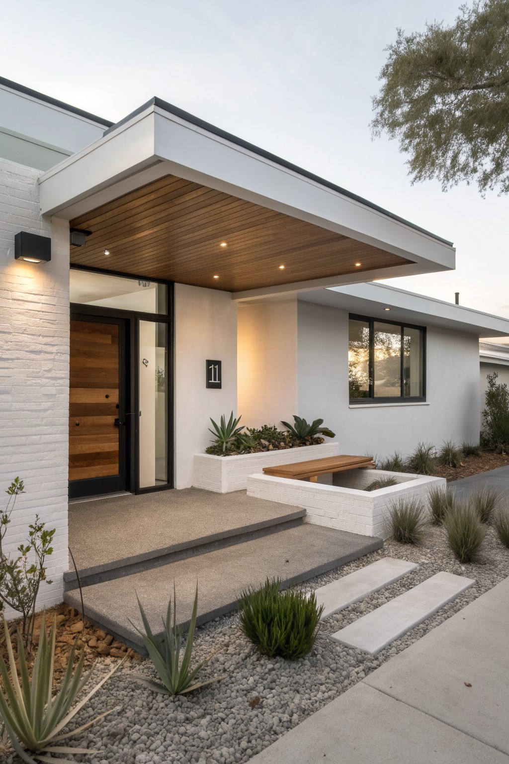 Modern white brick house exterior with cantilevered porch overhang featuring wooden slat ceiling and recessed lights, wood and glass entry door, potted succulents on bench, stone steps, and gravel landscaping.