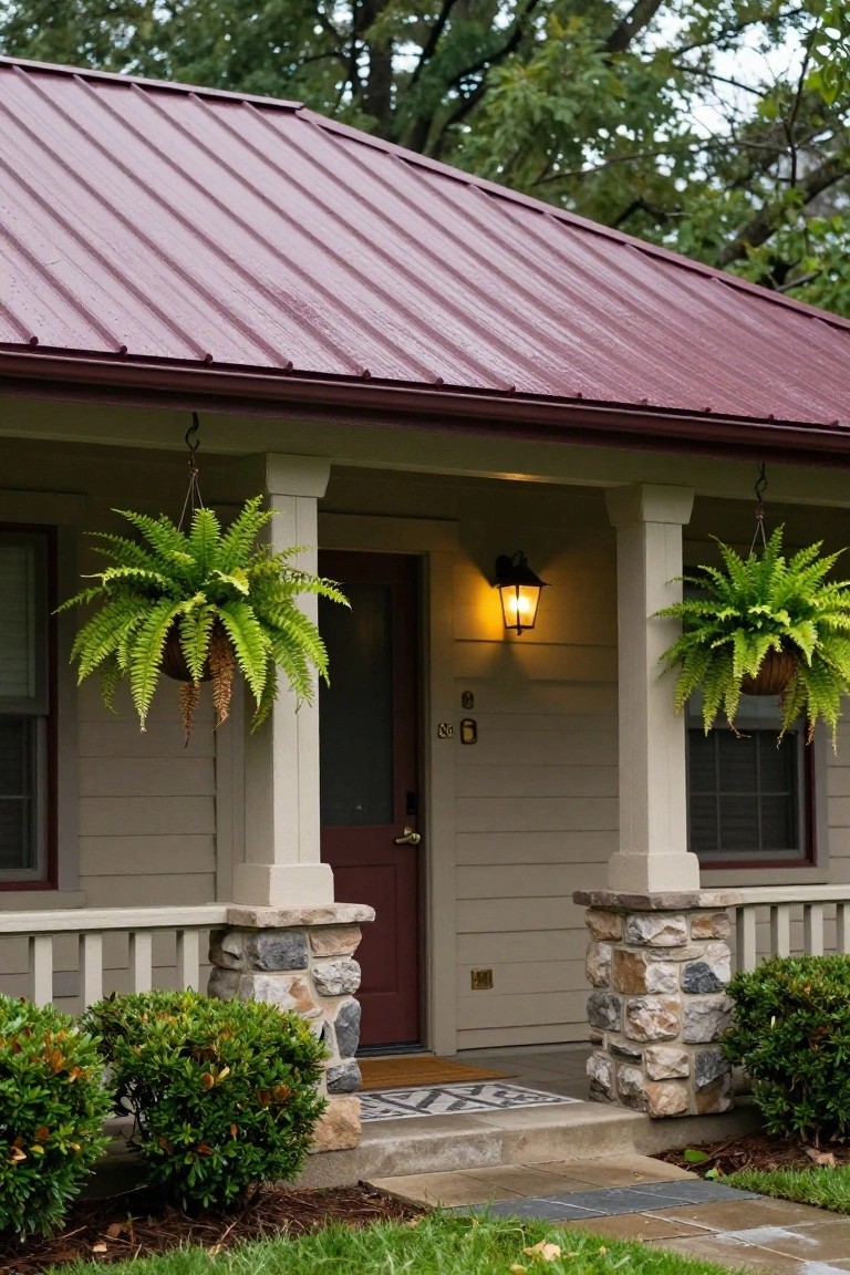 Front view of a single-story Craftsman-style house with maroon metal roof, beige horizontal siding, covered porch supported by stone pillars, red front door, hanging ferns, wall lantern, and boxwood shrubs beside concrete steps.