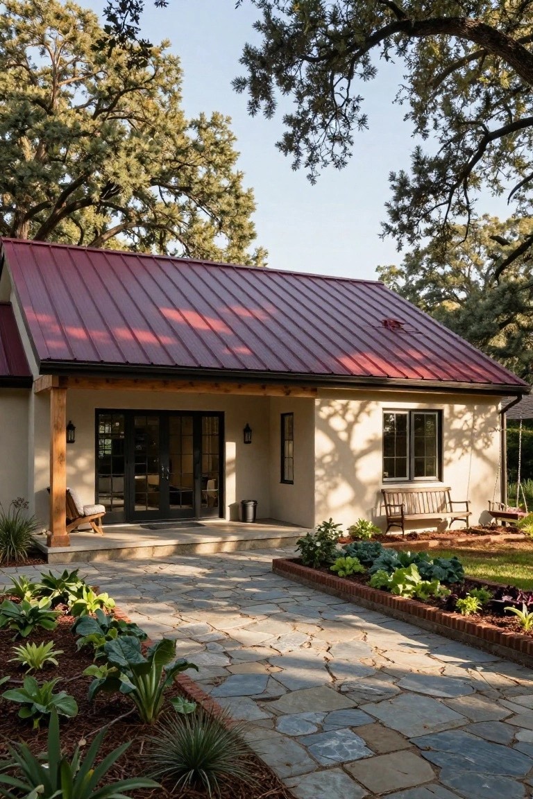 Small beige stucco house with maroon metal gable roof, wooden porch posts and bench, stone pathway, raised garden beds with vegetables, and oak trees in background.