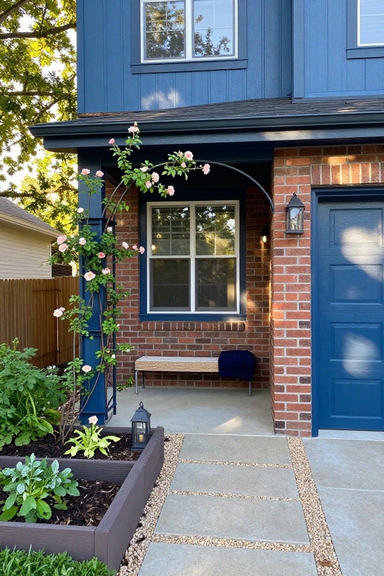 Front exterior of a blue clapboard house with red brick garage accents, tall metal arch covered in pink climbing roses, wooden bench on brick base, lantern light, raised garden bed with vegetables, and paver path edged in gravel.