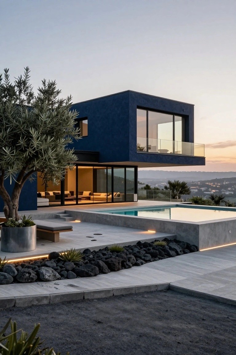 Modern deep blue rectangular house with cantilevered upper level, large glass windows, infinity-edge pool, concrete decking, rocks, plants, and olive tree overlooking a valley at dusk.