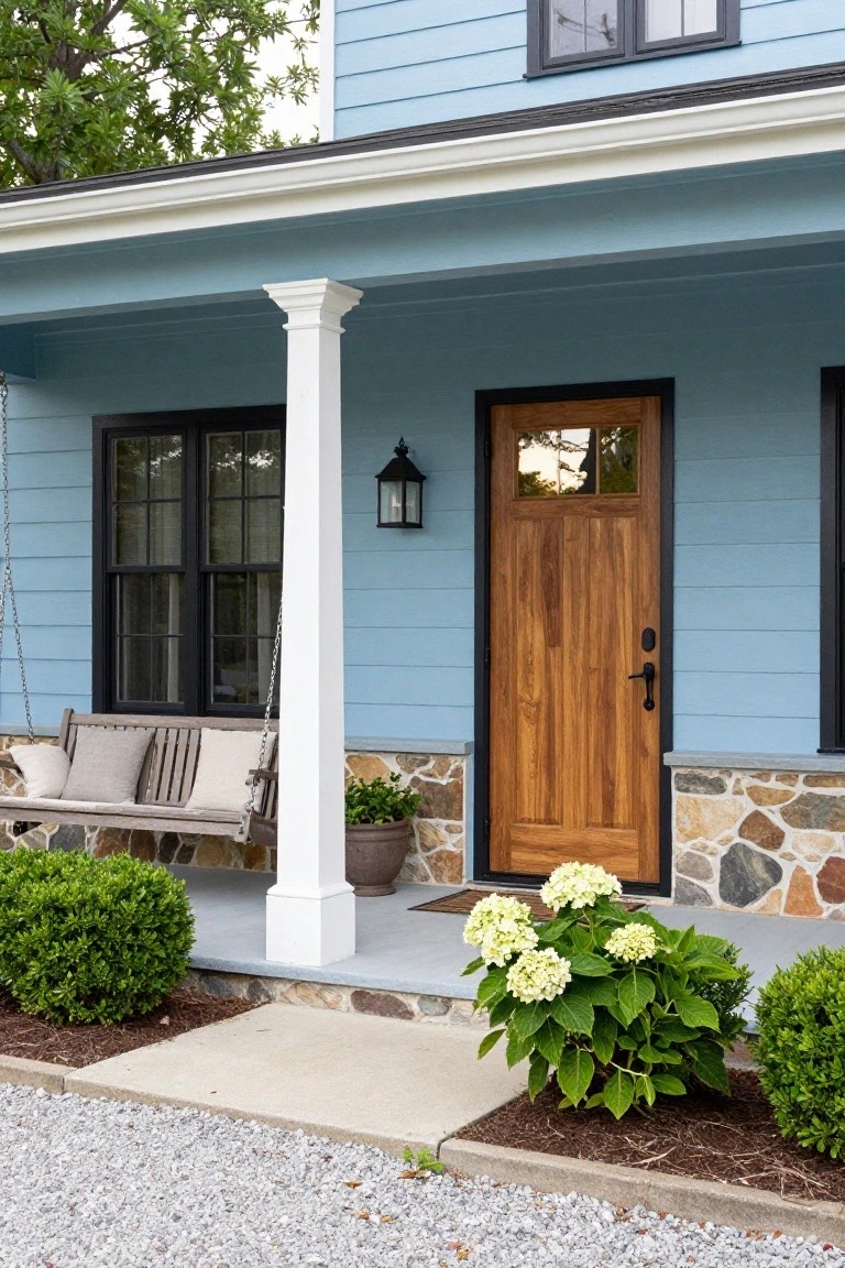 Light blue clapboard house exterior featuring a wooden front door with glass panels, white porch column supporting a hanging swing, black-framed windows, lantern light, stone foundation, and hydrangea plants by the steps.