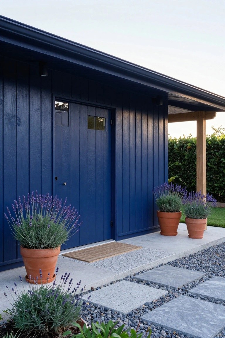 Dark blue painted vertical board-and-batten wooden shed with double doors and small window, flanked by three large terracotta pots of lavender, set against a gravel path with pavers and nearby hedge.