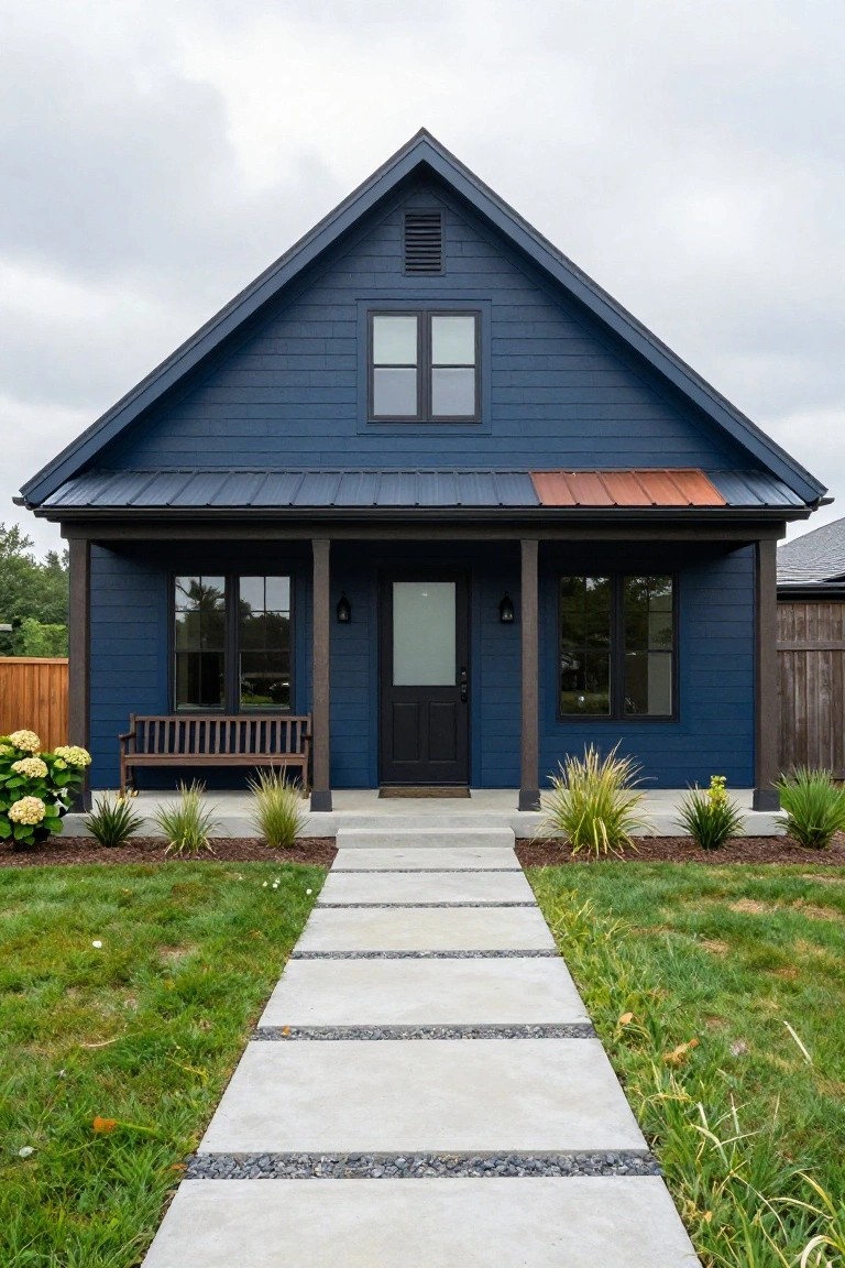 Front view of a two-story dark blue shingle-sided house with gabled roof, copper metal roof over the covered porch, black-framed windows and door, wooden bench on porch, concrete paver path lined with grasses and hydrangeas in a grassy yard.