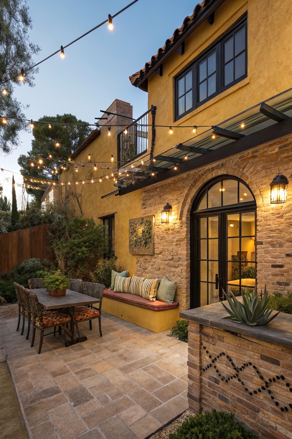 Backyard patio featuring a wooden table with upholstered chairs, built-in yellow bench with cushions against ochre stucco wall with brick archway door, overhead string lights, potted plants, and stone pavers at dusk.