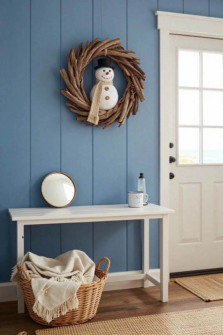 Blue shiplap wall in a coastal entryway featuring a driftwood snowman wreath above a white console table with round mirror, enamel mug, glass lantern, and wicker basket of blankets next to a white door with glass panels overlooking the ocean.