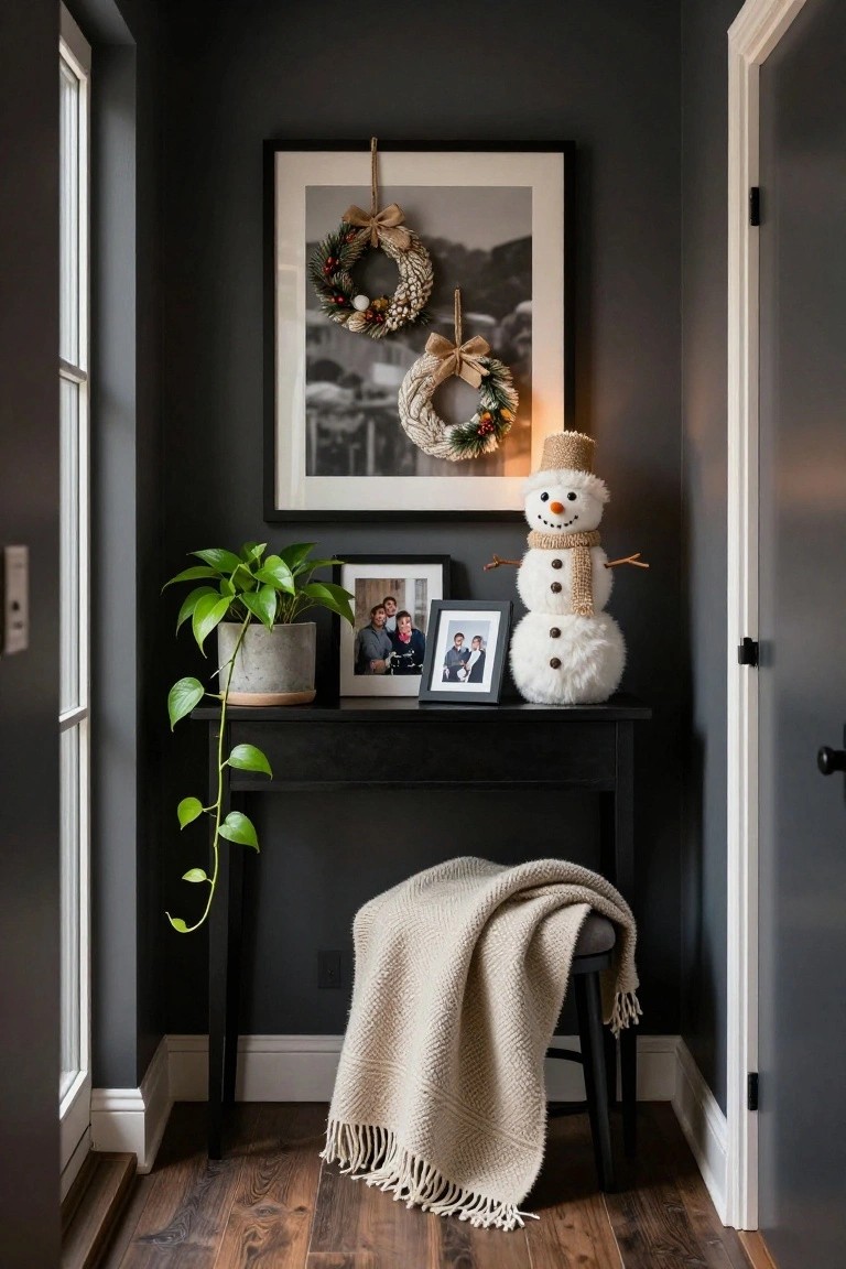 Narrow indoor nook with dark gray walls, a black console table holding a white plush snowman figure, potted green plant, family photos in frames, and a beige throw-draped stool nearby, with wreaths and a large framed photo on the wall.