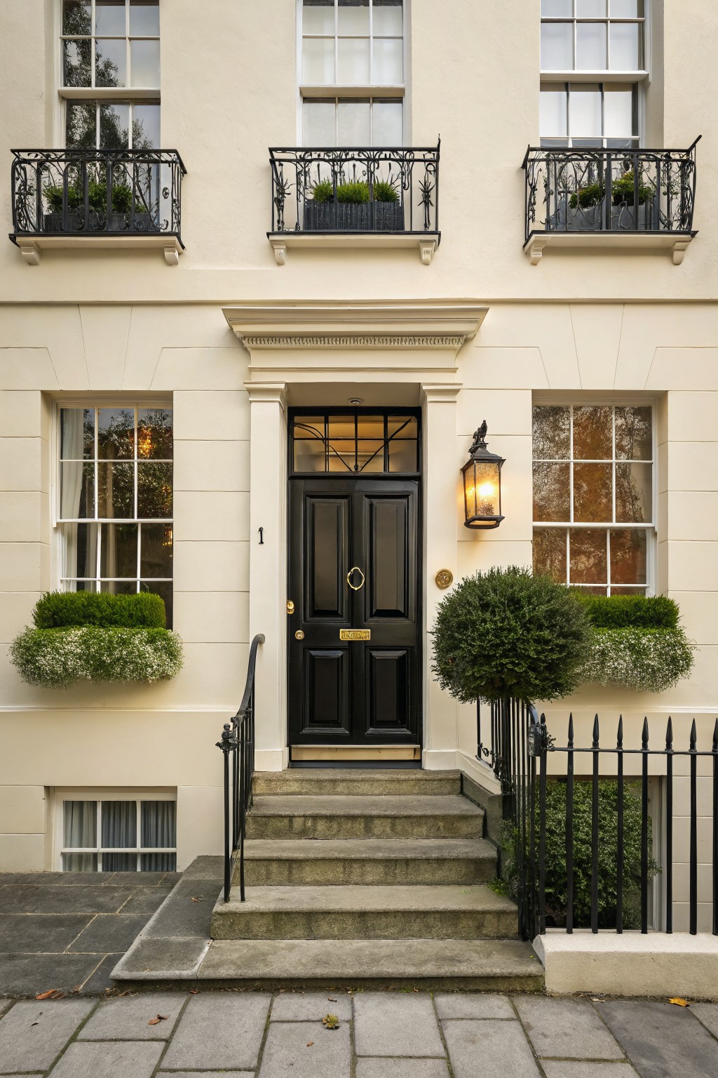 A three-story Georgian-style townhouse with soft off-white stucco walls, glossy black front door numbered 1, sash windows with green boxwood plantings, wrought iron balconies and railings with planters, stone steps, and a lantern light.
