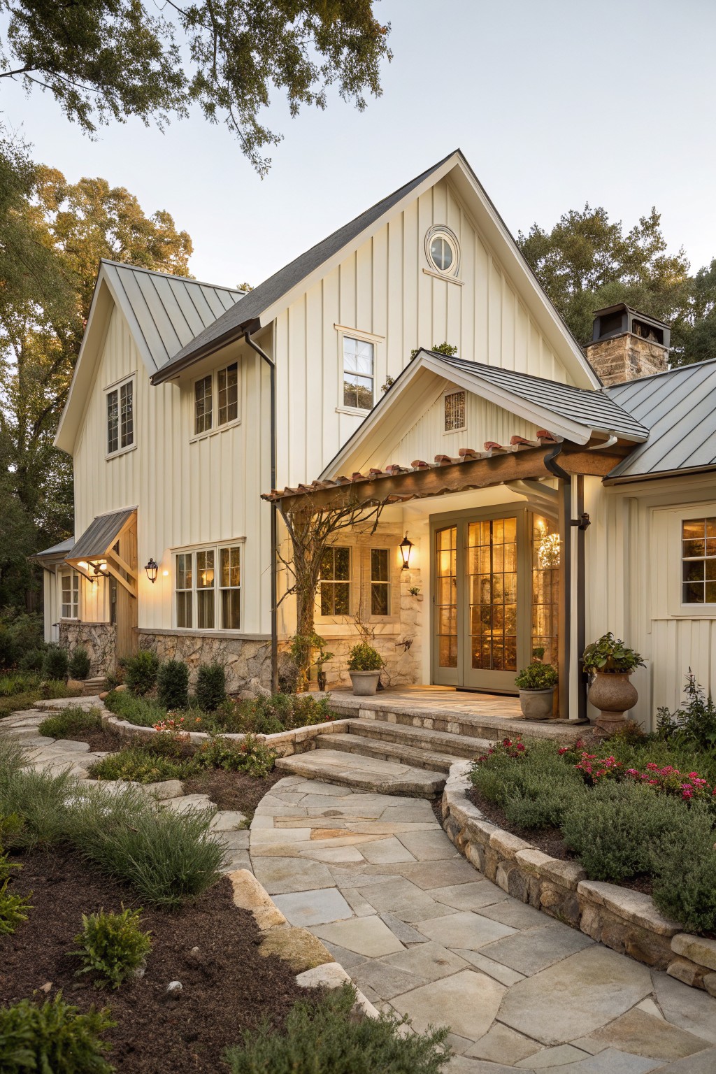 Two-story house exterior with cream board-and-batten siding, gray metal roof, stone foundation and accents, wooden pergola over glass entry doors, and curved stone pathway with shrubs and plants.