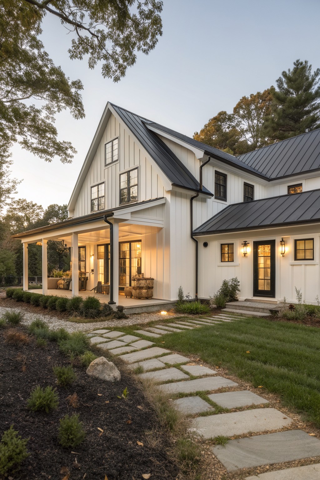 Two-story house exterior with soft off-white board and batten siding, black metal gable roof, wraparound porch with black posts and glass doors, stone pathway, landscaping beds, and surrounding trees at dusk.