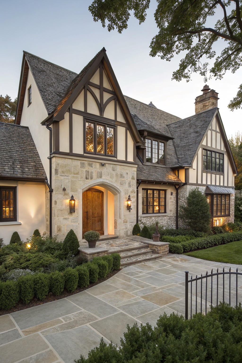 Two-story Tudor-style house exterior with beige stucco walls, dark timber framing, stone-clad arched entry door, slate roof, lanterns, and landscaped front yard with shrubs and paver path at dusk.