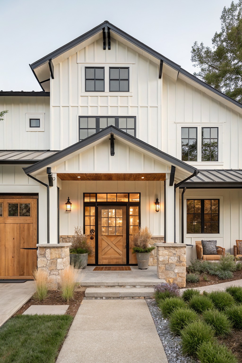 Front view of a two-story house featuring soft white board-and-batten siding, black metal roof and trim, wooden garage door, stone pillars supporting a covered porch, and a wooden entry door with sidelights and lanterns.
