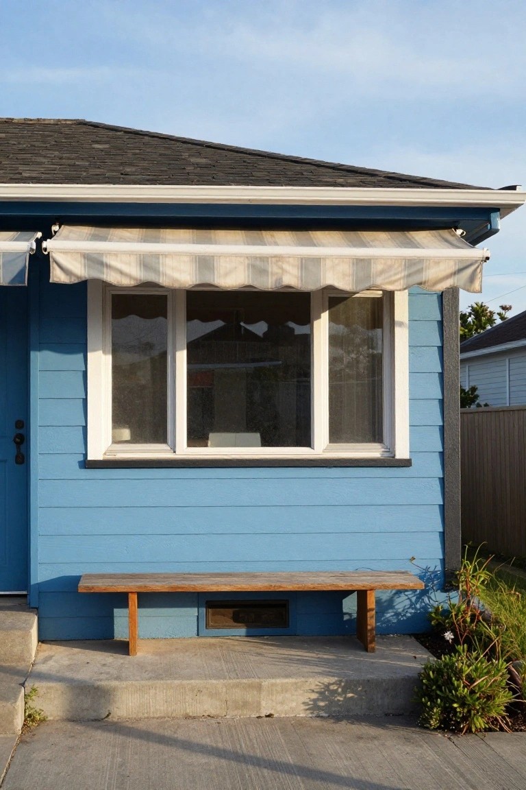 Blue clapboard house exterior featuring a white-framed triple window under a beige striped awning, wooden bench on concrete steps, small shrubs, and a side door.