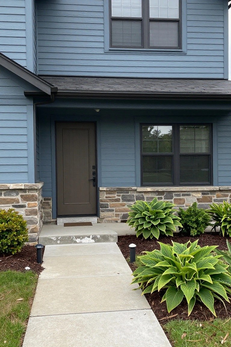 Front view of a two-story house with light blue-gray horizontal siding, dark wood entry door under a small covered porch flanked by stone pillars, hosta plants in mulch beds, and a concrete walkway.