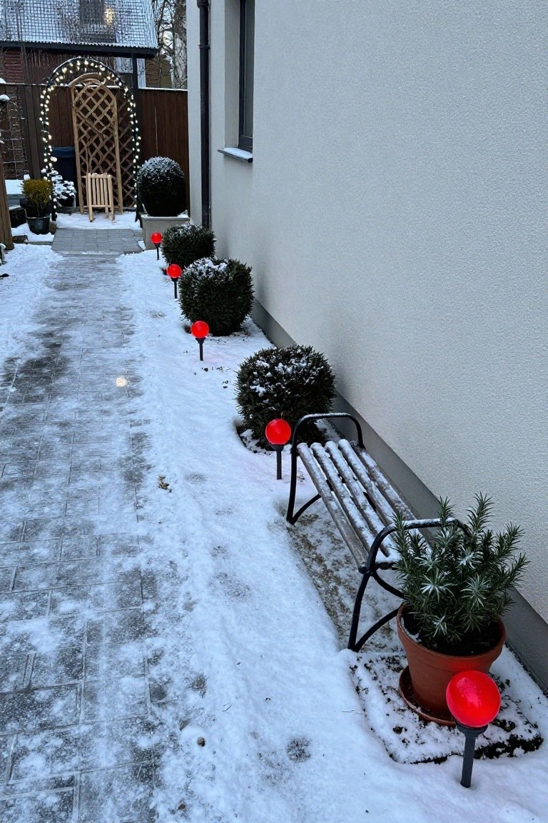 Snowy paved pathway along a white house wall lined with red spherical lights on black stakes next to snow-dusted boxwood shrubs, a metal bench, potted rosemary plant, and red lights by a wooden archway in the background.