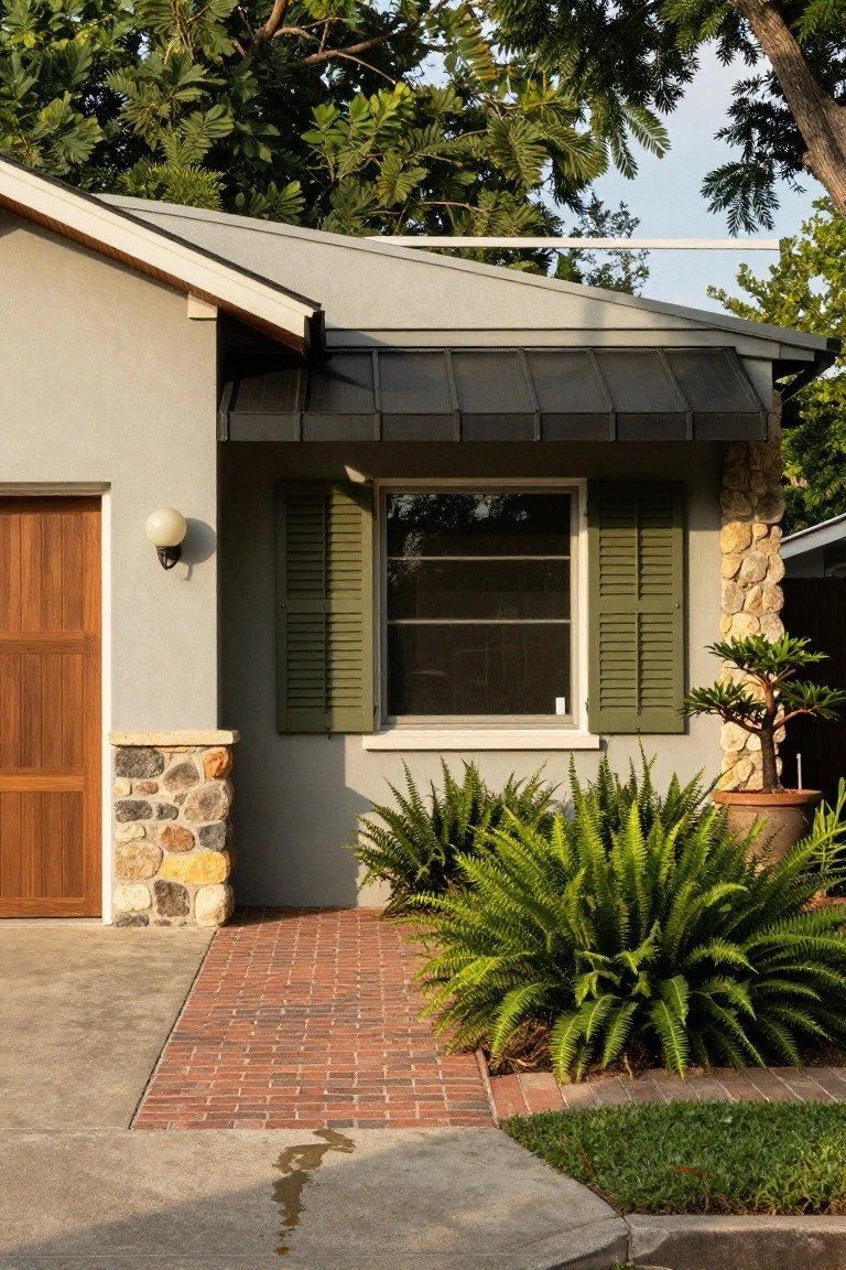 Front exterior of a house featuring light gray stucco walls, wooden garage door, stone pillar at corner, window with green shutters and dark awning, potted palm tree, ferns, brick pathway, and concrete driveway.