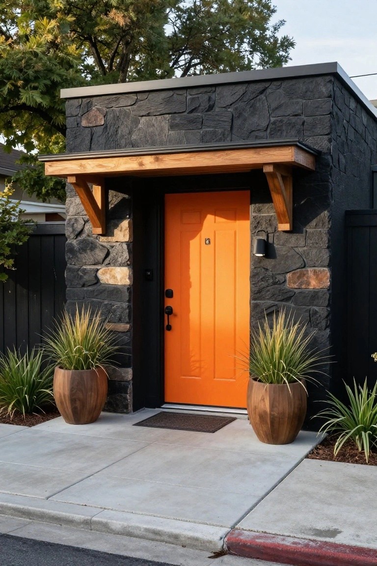 Black stone house exterior with glossy orange front door under wooden awning, flanked by two large wooden pots of tall ornamental grasses on a concrete walkway.
