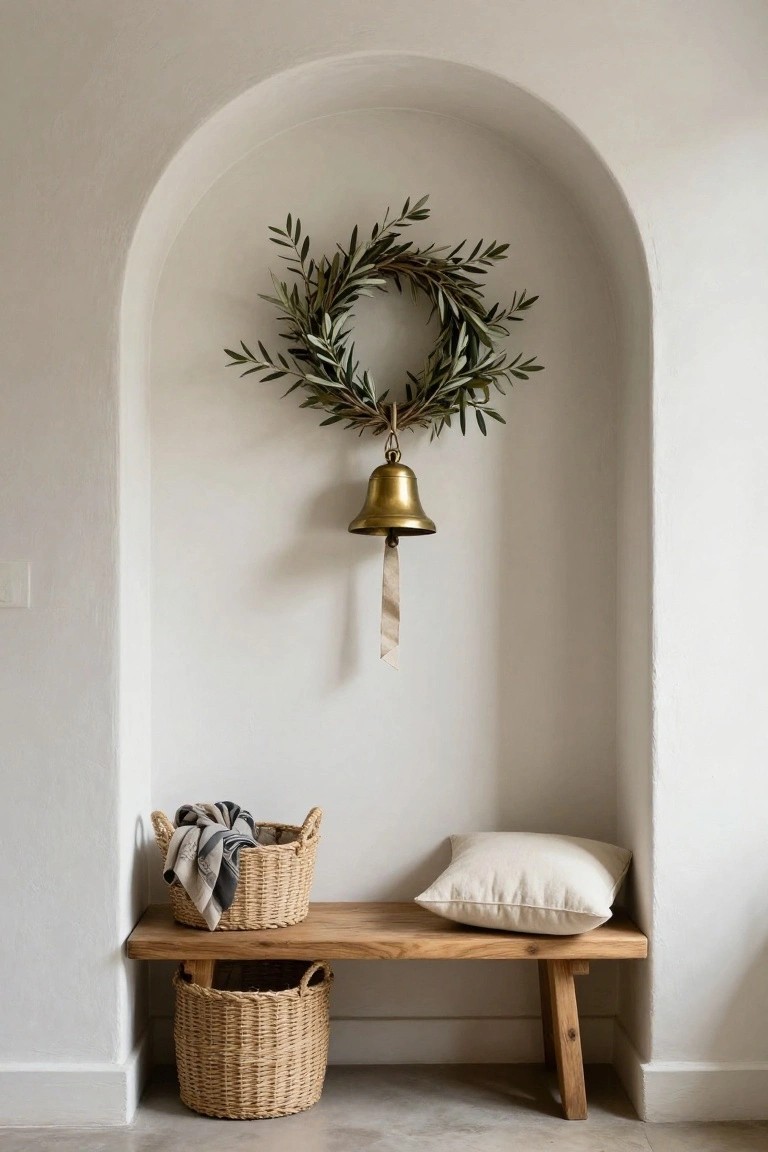 White arched wall niche with olive leaf wreath centered above a hanging brass bell on beige ribbon, wooden bench below with white pillow, two wicker baskets, and draped linens.