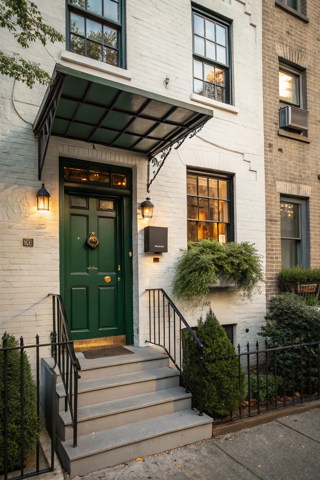 White brick townhouse facade with dark green paneled front door, brass knocker and handle, black metal awning and railing, lanterns, steps, and boxwood shrubs on a city sidewalk.