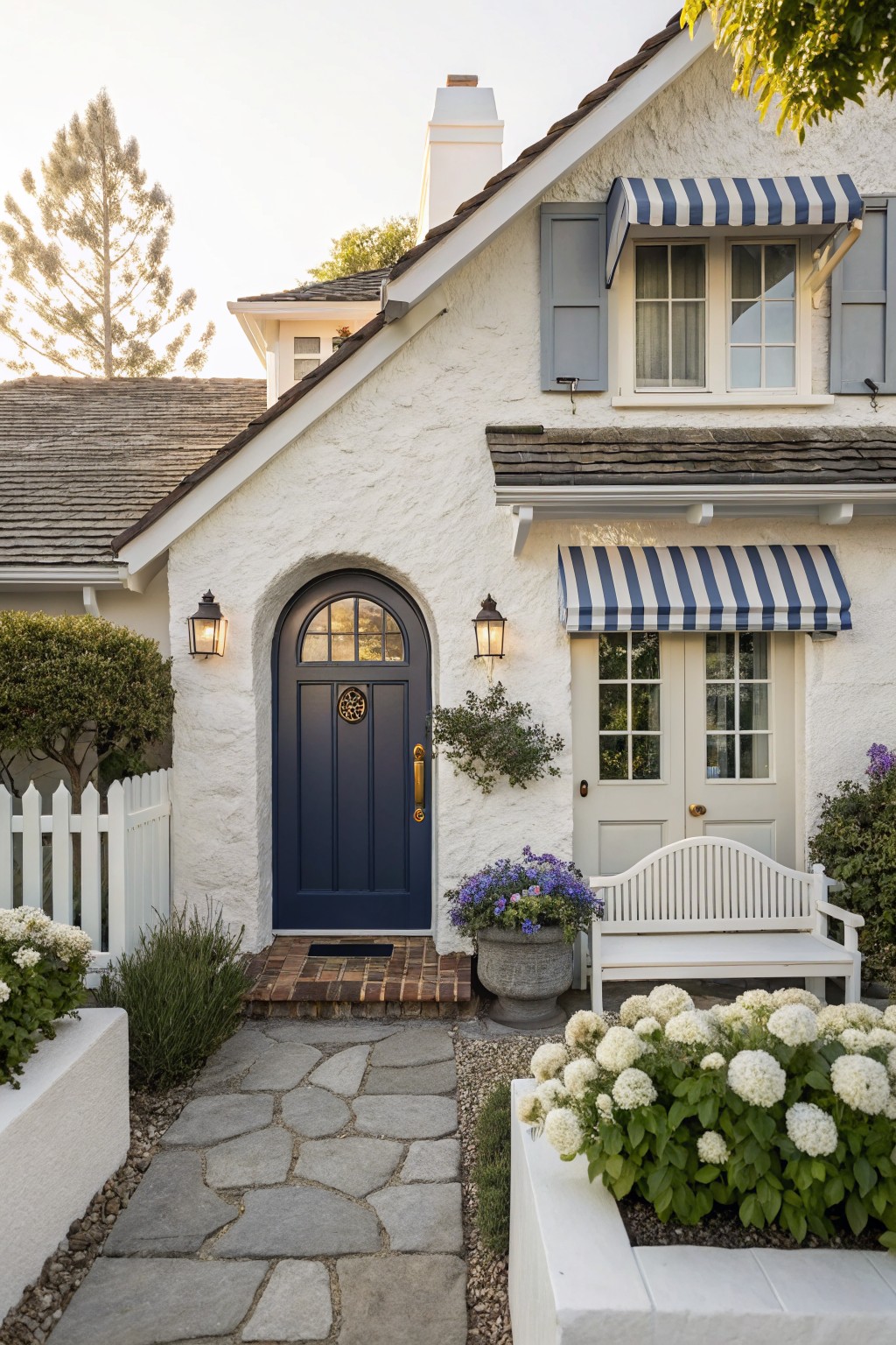 White stucco house exterior featuring a navy blue arched entry door with gold knocker, flanked by lanterns, a stone pathway, white picket fence, and hydrangea plantings.