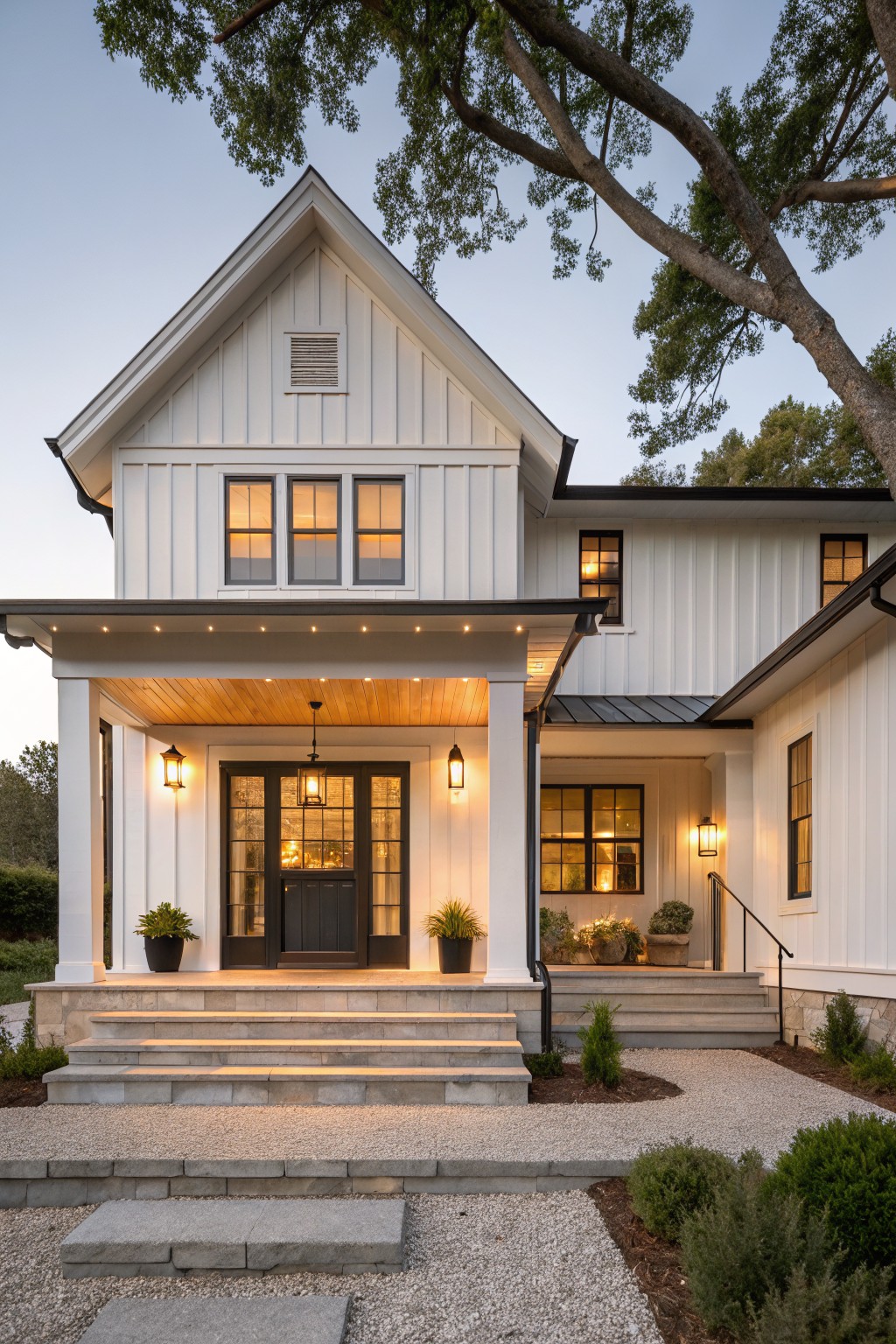 White board-and-batten sided house exterior with covered front porch, wood plank ceiling, central hanging lantern, flanking wall lanterns, dark double doors, stone steps, gravel path, potted plants, shrubs, and overhanging tree branches at dusk.