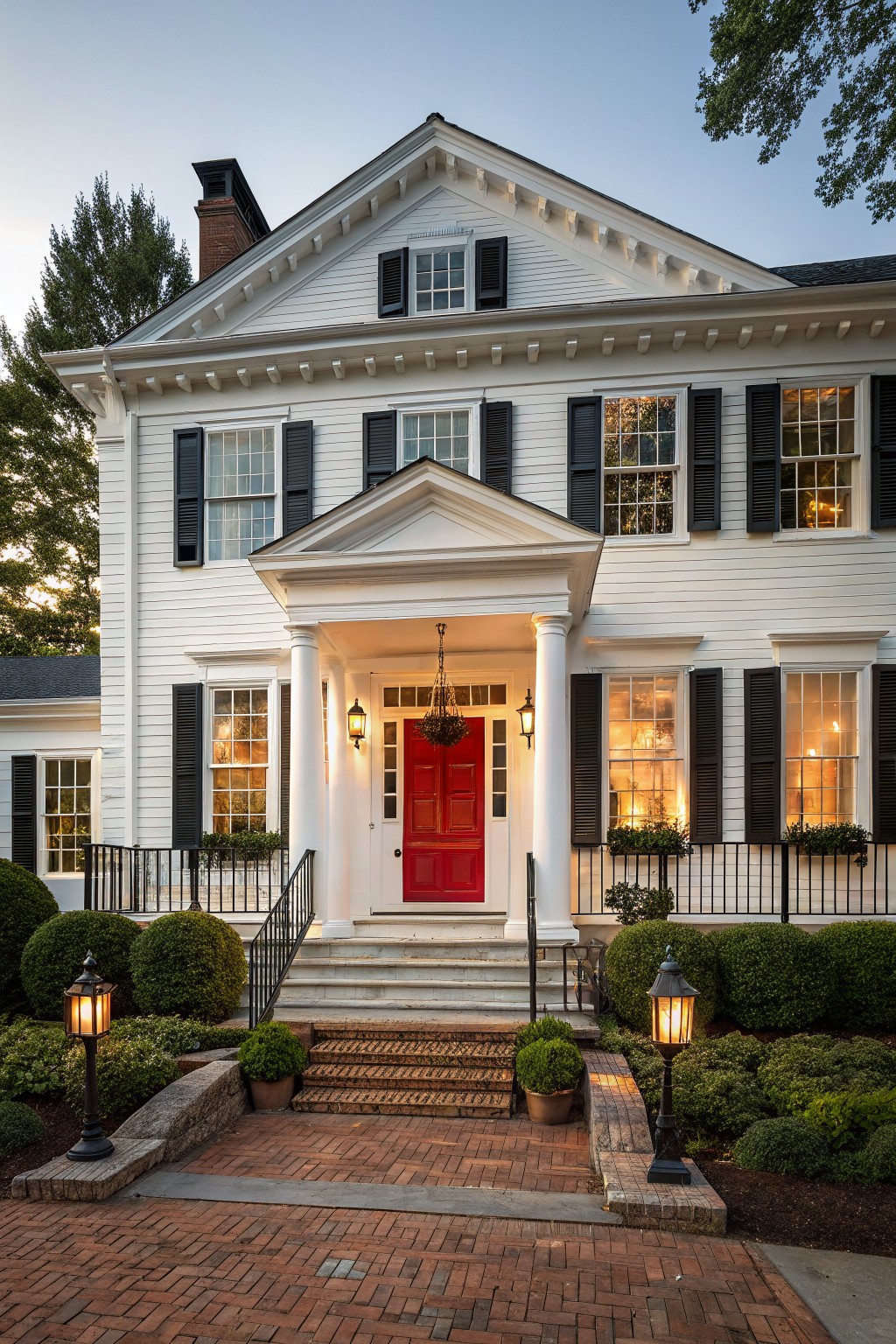 White colonial-style house exterior with black shutters, red front door under a portico with columns, brick steps, iron railings, lanterns, and landscaped shrubs at dusk.