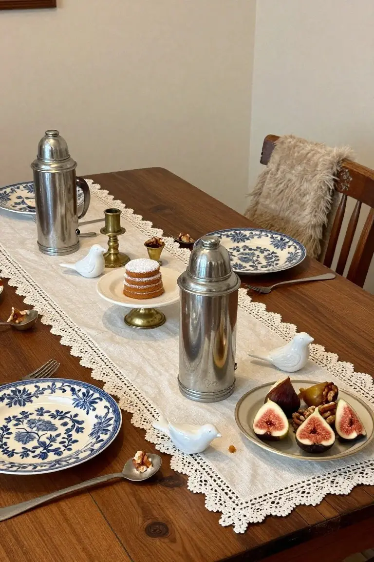 Wooden dining table decorated with white lace runner, two tall silver flasks, small white porcelain birds, blue floral plates, figs with nuts, and miniature cakes on a stand.