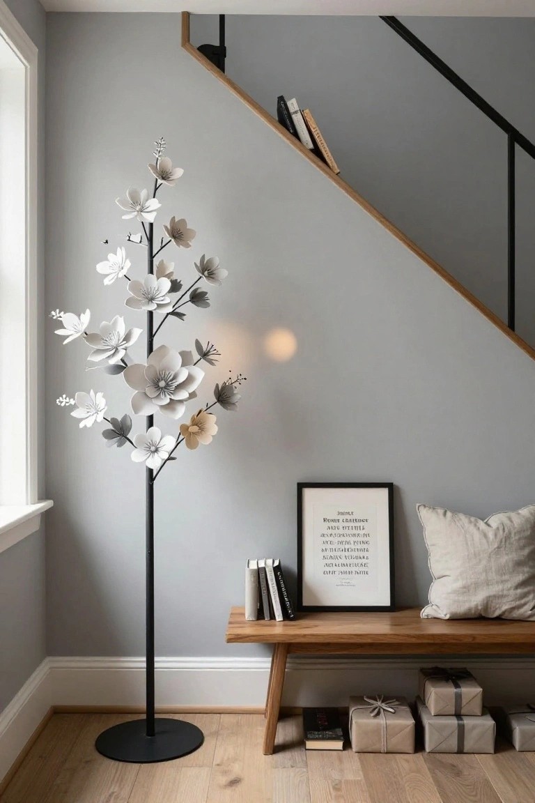 Tall floor lamp shaped like a tree with layered white flowers, positioned in a gray-walled interior corner near a wooden bench with pillows, books, framed art, and wrapped gifts, with a staircase visible.