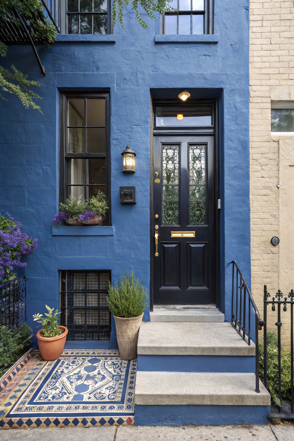 Navy blue painted brick townhouse exterior featuring a black double front door with glass panels, brass hardware, potted plants on steps, purple flowers in window box, and blue-white patterned rug at entry next to beige brick building.