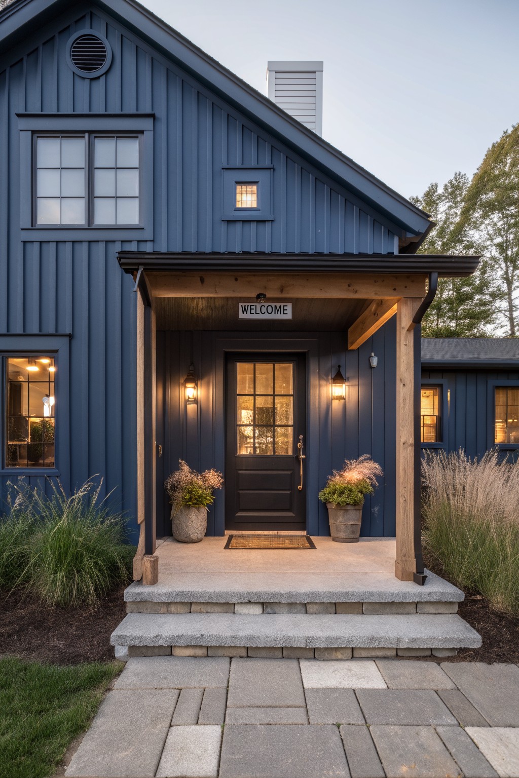 Navy blue board-and-batten house exterior with a covered wooden porch over a black glass-paneled front door, flanked by lanterns and potted plants, stone steps, and ornamental grasses.