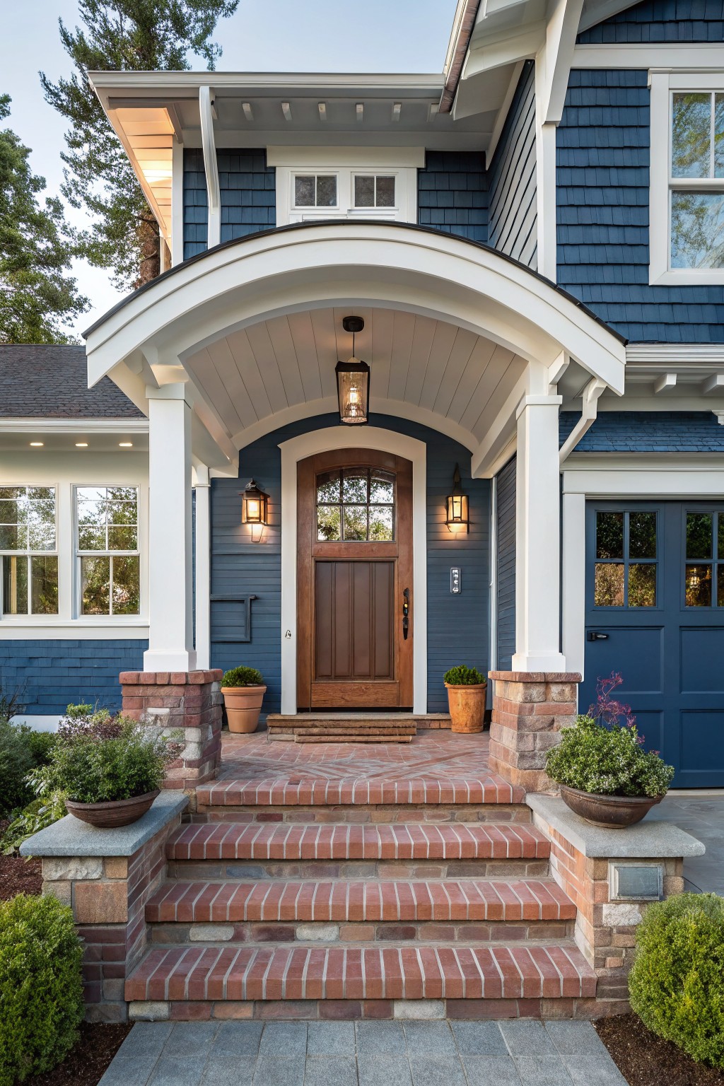 Front view of a two-story navy blue shingle house with white arched portico supported by columns, dark wood arched door with glass panels, brick steps, potted plants, and landscaped entryway.