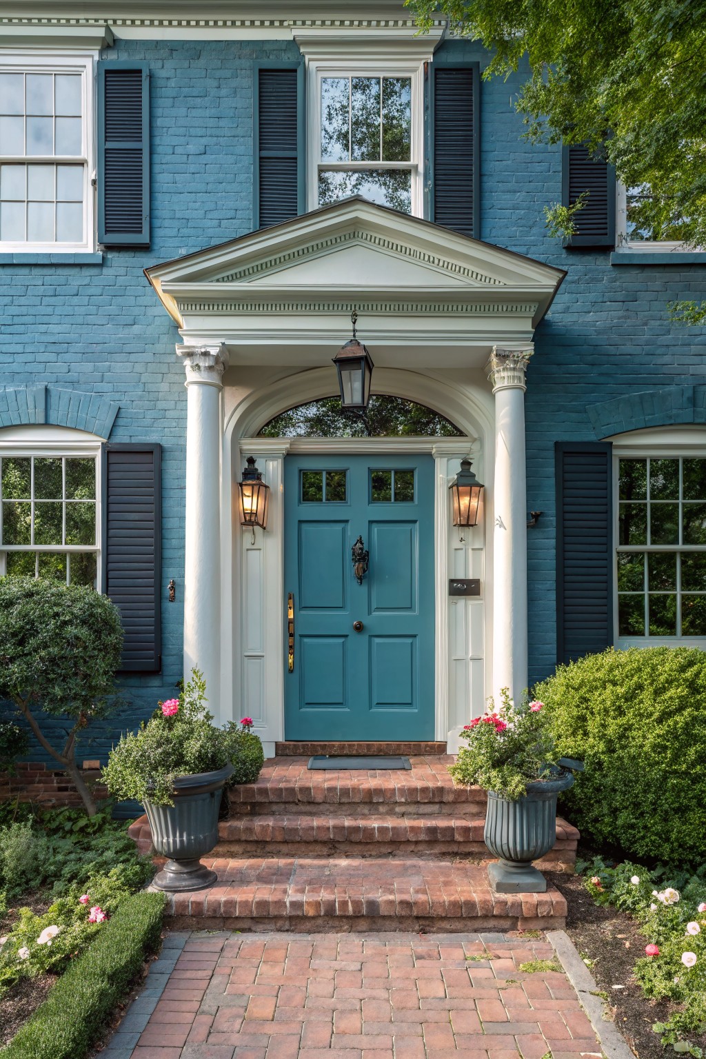 Blue brick house facade with teal front door centered under white pedimented portico supported by columns, flanked by black lanterns, brick steps, potted plants, and brick walkway.