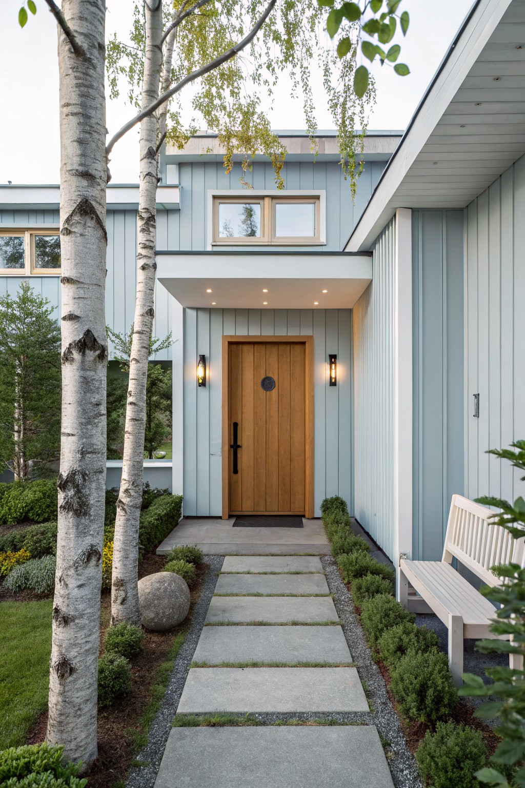 Front exterior of a light blue vertical board-and-batten house with a large round-topped wooden door centered under a covered entry, flanked by black wall lights, leading to a concrete paver pathway lined with shrubs, birch trees, a boulder, and a white bench.