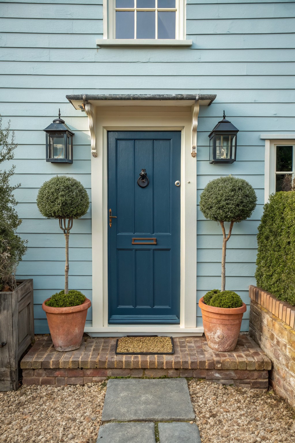 Pale blue clapboard house exterior with navy paneled front door, flanked by two tall potted topiary trees in terracotta pots, lanterns on white trim, brick steps, and gravel path.