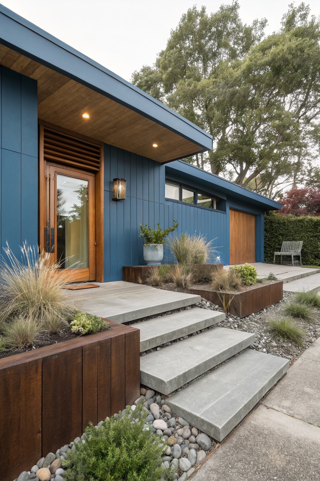 Blue vertical board-and-batten house exterior with wood front door, single-car garage, concrete steps to entrance flanked by raised wooden planters containing grasses and low plants, pebble ground cover, and nearby shrubs.
