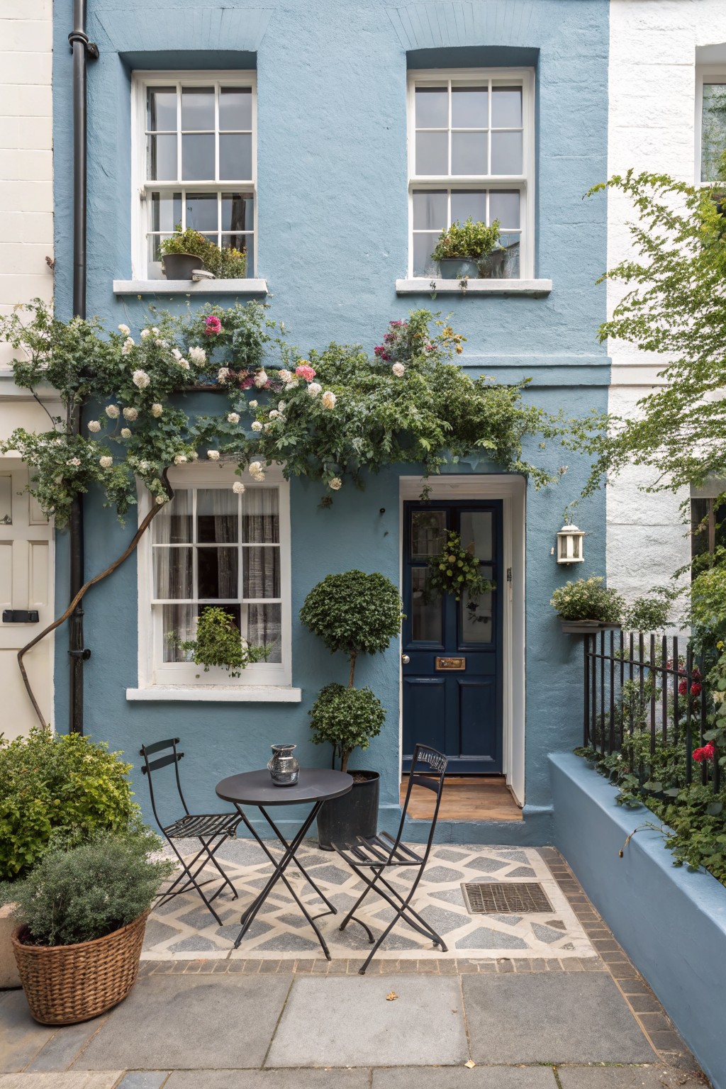 Pale blue terraced house exterior with white sash windows, navy front door, climbing white roses over the doorway, potted plants including topiary balls and a small metal bistro table with chairs on a tiled front patio.