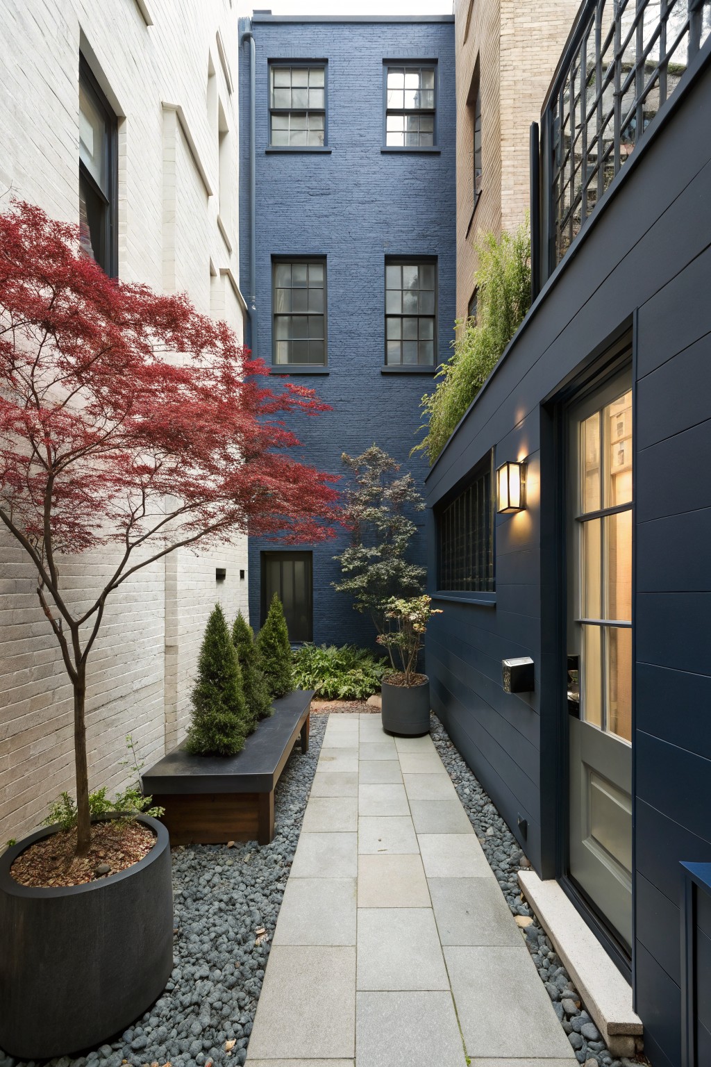 Narrow stone pathway with gravel edging, potted plants, a wooden bench, and a tall red Japanese maple tree leading to a glass door on a dark blue paneled wall between brick buildings.