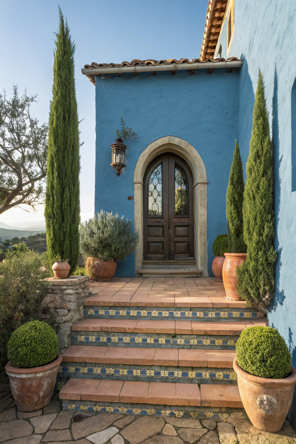 Blue stucco house exterior with arched wooden double door, flanked by two tall slender cypress trees, terracotta pots with shrubs and topiaries on colorful tiled steps leading to the entrance.