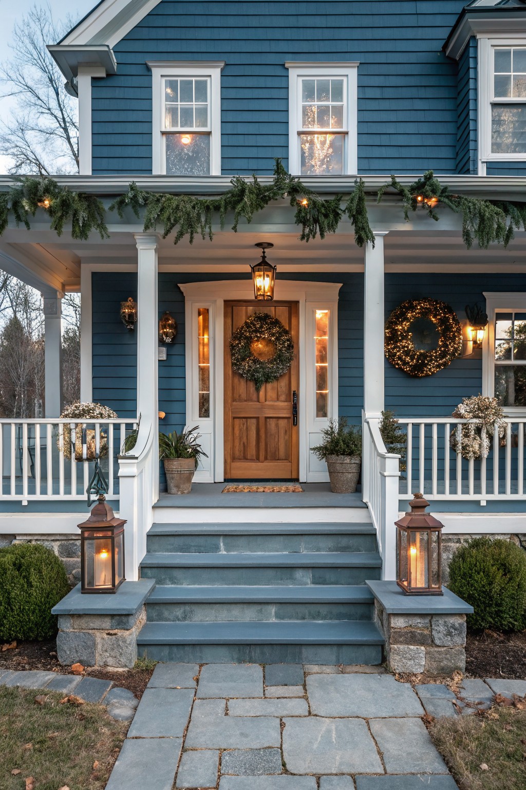 Blue shingle-style house exterior with Christmas decorations including evergreen garlands on the porch roof and columns, wreaths on the door and porch supports, lit lanterns on steps, and potted plants beside bluestone stairs.