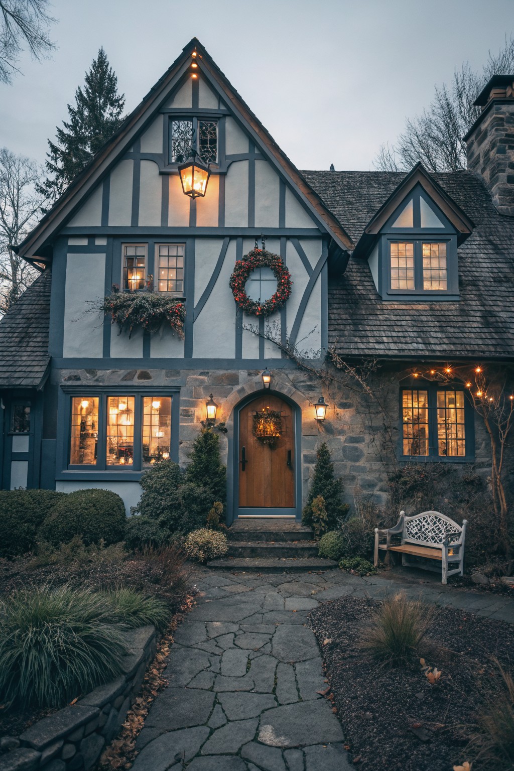 Twilight photo of a two-story blue Tudor-style house exterior with Christmas wreaths on door and windows, lanterns flanking arched entry, string lights on eaves and plants, potted trees, and stone pathway.