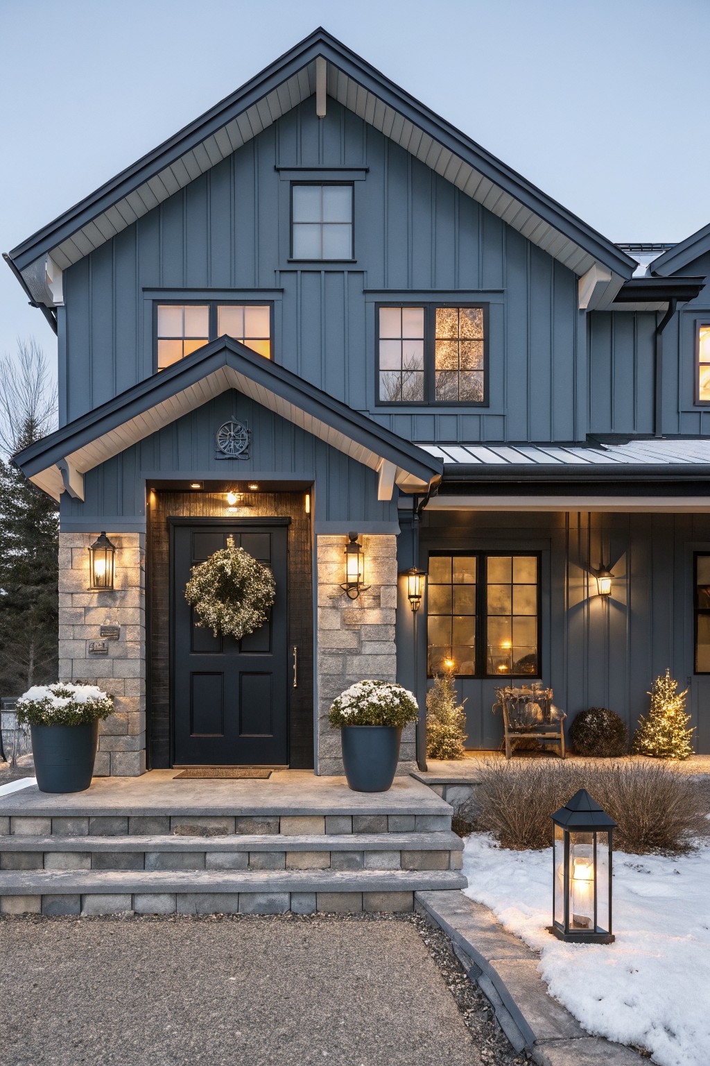 Blue vertical board-and-batten house exterior at dusk with snow, black front door decorated with white Christmas wreath, flanked by stone pillars with hanging lanterns and blue pots of plants, wooden chair, small Christmas trees, and yard lantern.