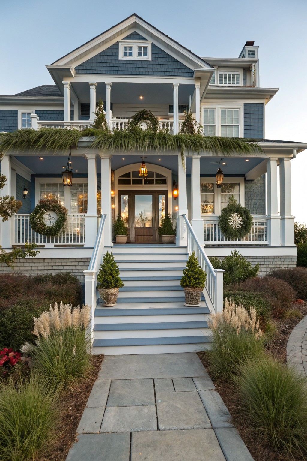 Blue shingle-style house with white porch and columns, decorated for Christmas with evergreen wreaths on railings and balcony, garlands overhead, lanterns, and potted trees beside blue steps leading to the entry.