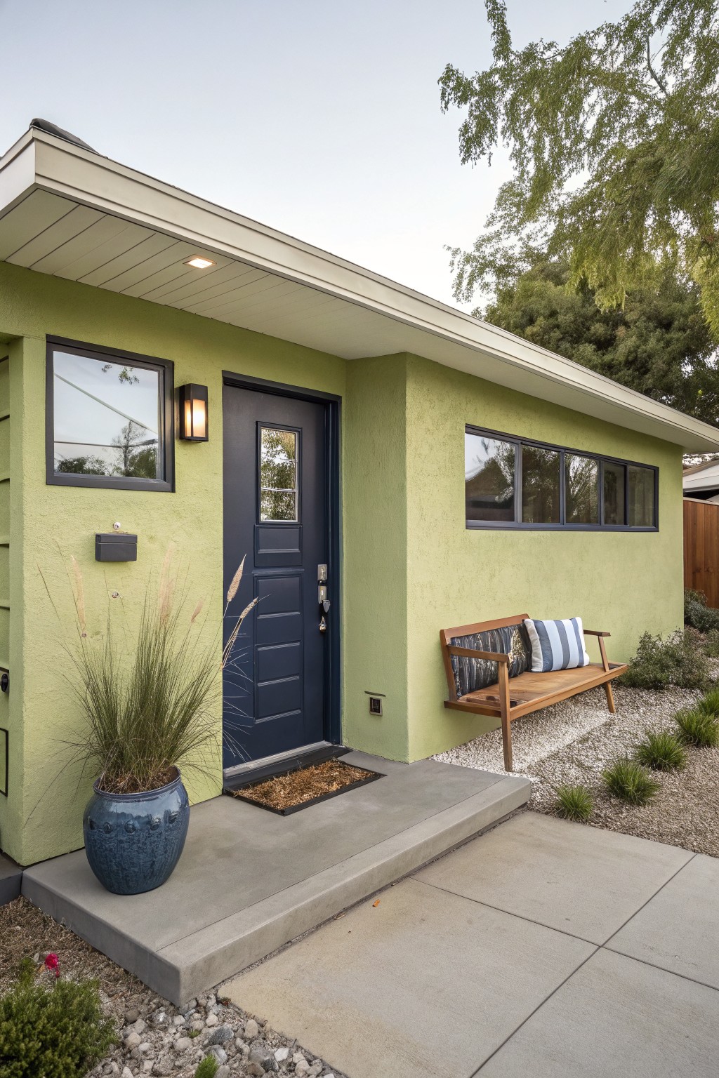 Light green stucco house exterior featuring a navy blue front door with sidelight, wall-mounted light, pampas grass in blue pot, wooden bench with cushions, and gravel landscaping.