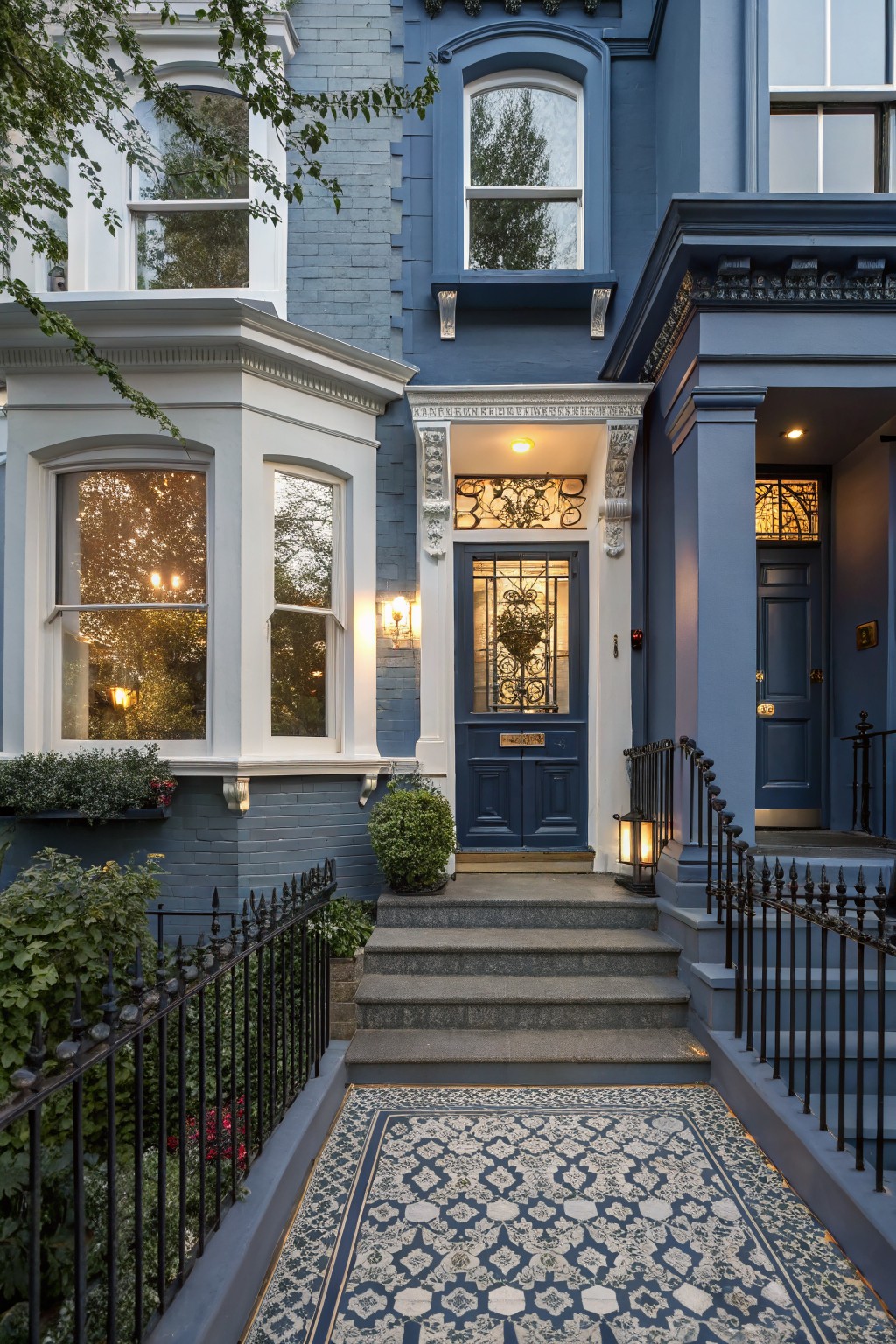 Blue Victorian-style townhouse facade with white trim around bay windows and doors, stained glass panels, wall lanterns, black wrought iron railing, potted plants, and a blue patterned rug on the stone steps.