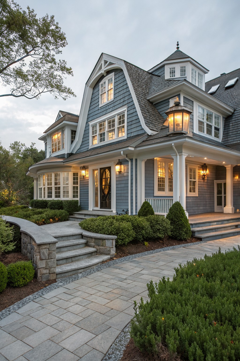 A two-story blue shingle-style house with white trim, wraparound porch, multiple lit lanterns, stone entry steps, and boxwood shrubs along a curved paver pathway.