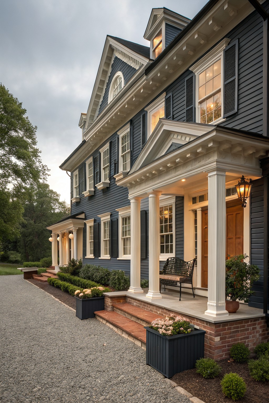 A two-story navy blue clapboard house with white trim, classical columns supporting a pedimented porch, double front doors, potted plants, and landscaping along a gravel driveway and brick steps.